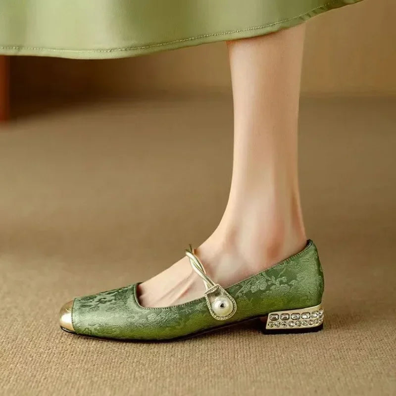 A woman wearing green low-heel Mary Janes with a twisted gold strap, gilded cap toe and a crystal-set heel