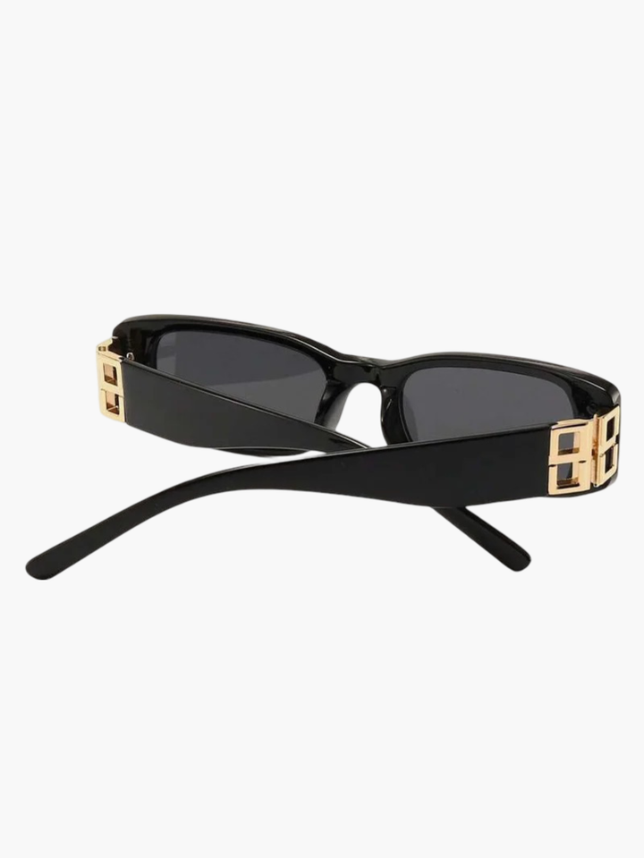 A woman smiling outdoors wearing slim black rectangular sunglasses with gold-tone temple detail.