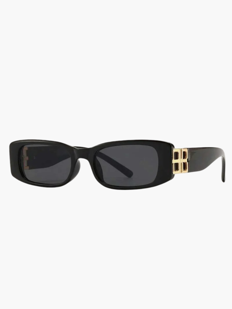 A woman smiling outdoors wearing slim black rectangular sunglasses with gold-tone temple detail.