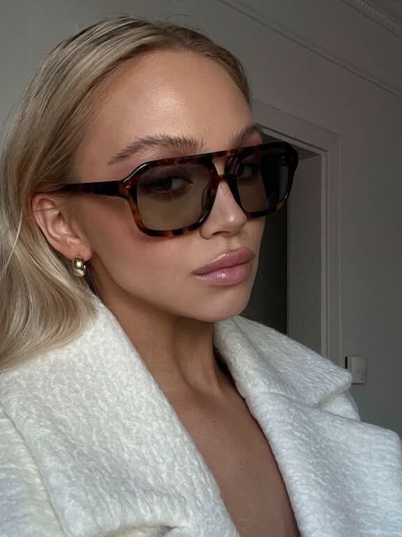 A woman wearing oversized tortoiseshell aviator sunglasses with green-tinted lenses on a sunny day outdoors.