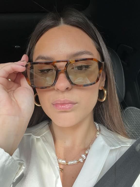 A woman wearing oversized tortoiseshell aviator sunglasses with green-tinted lenses on a sunny day outdoors.