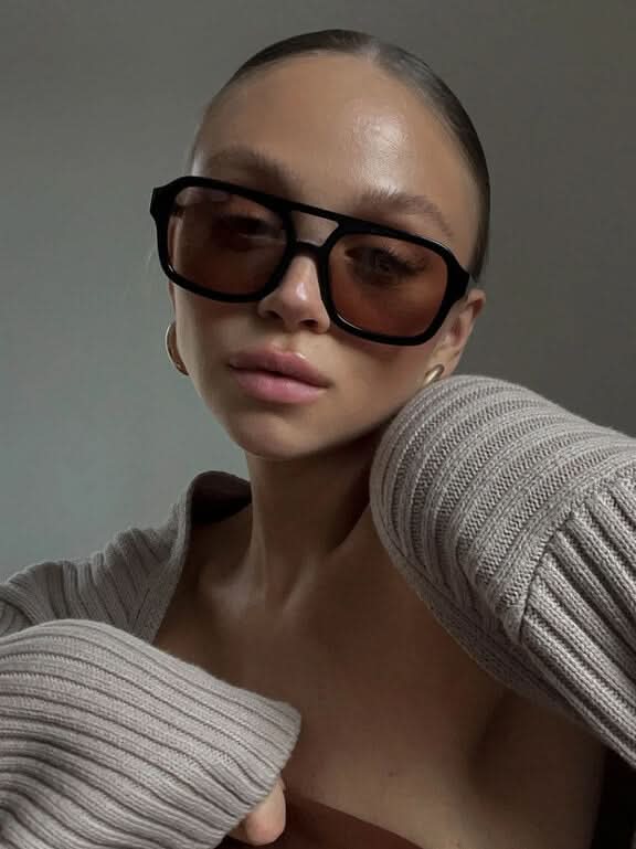 A woman wearing oversized tortoiseshell aviator sunglasses with green-tinted lenses on a sunny day outdoors.