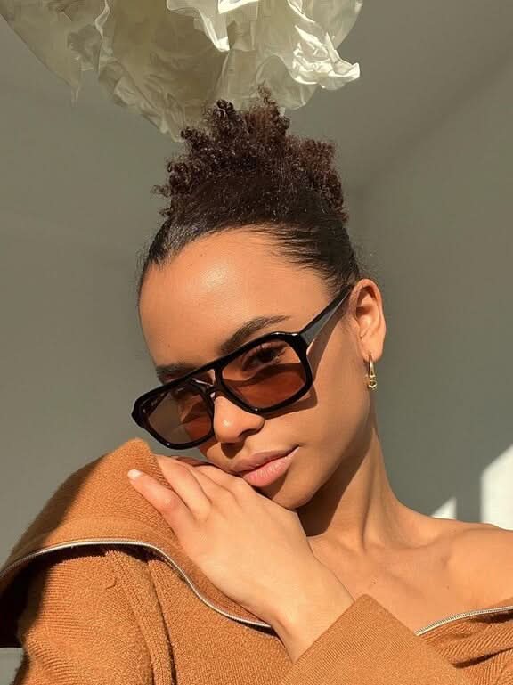 A woman wearing oversized tortoiseshell aviator sunglasses with green-tinted lenses on a sunny day outdoors.