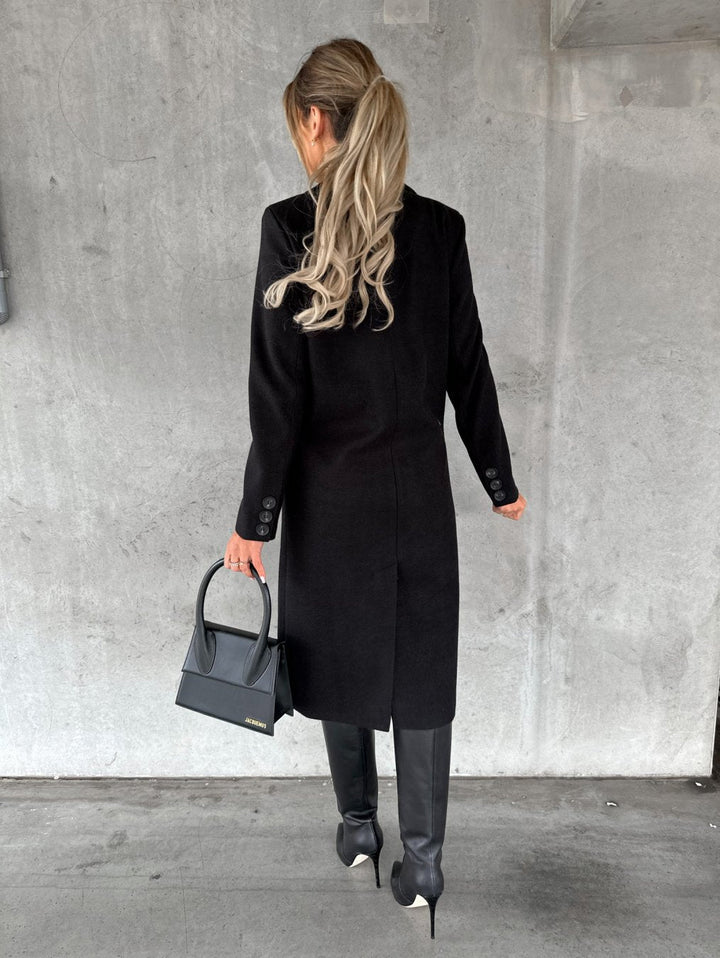 A woman wearing a long waisted coat, standing elegantly outdoors