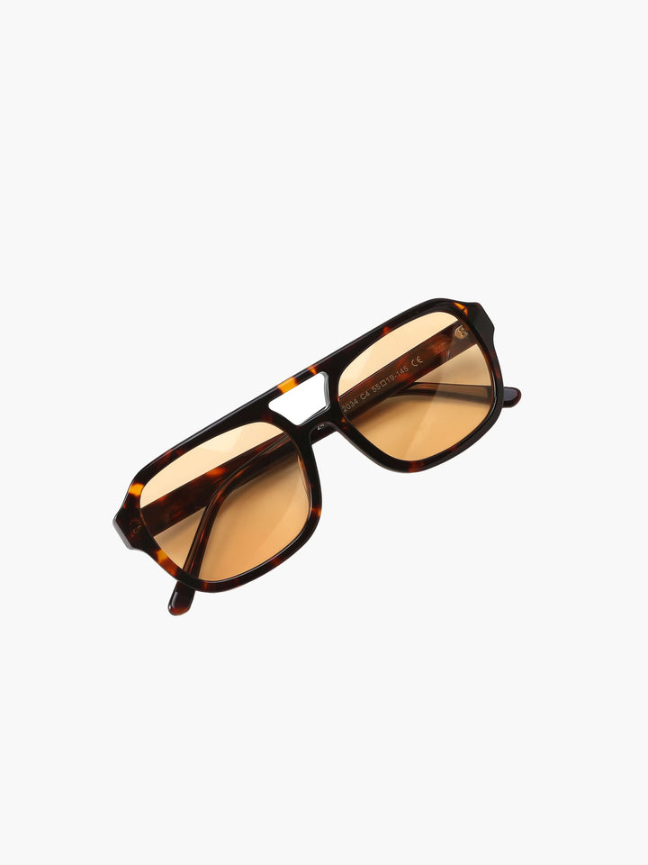 A woman wearing oversized tortoiseshell aviator sunglasses with green-tinted lenses on a sunny day outdoors.