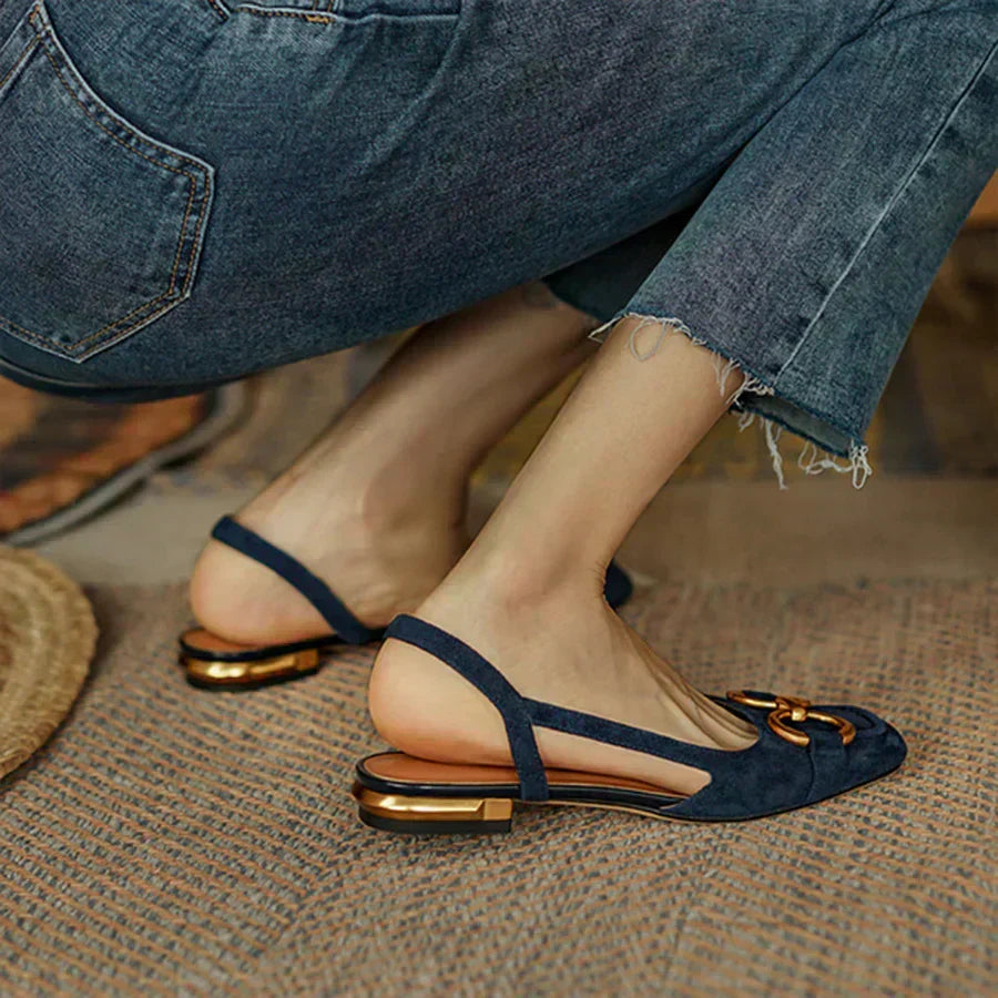 A woman wearing navy square-toe open-back flats with a small golden buckle detail.