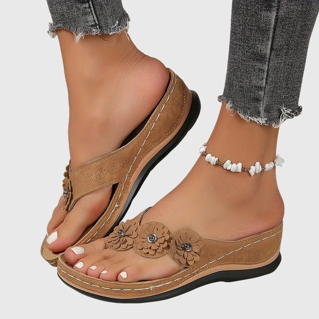 A woman wearing tan flower-trim flip-flops with a toe-post front and a low wedge sole.