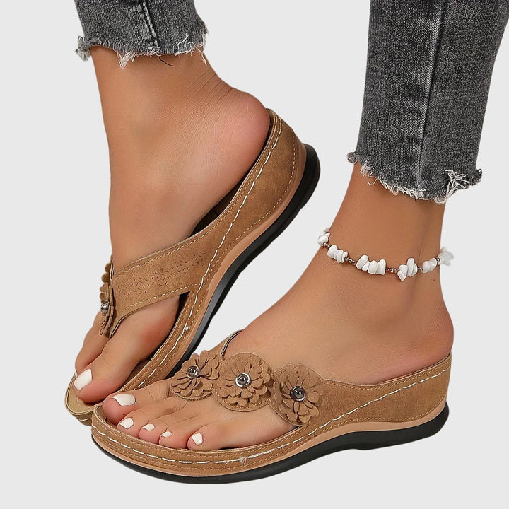A woman wearing tan flower-trim flip-flops with a toe-post front and a low wedge sole.