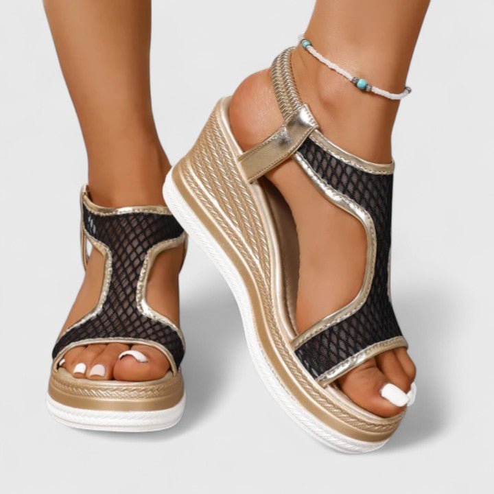 A woman wearing golden platform wedge sandals with a T-bar front, open-weave panels and a stretchy slingback.