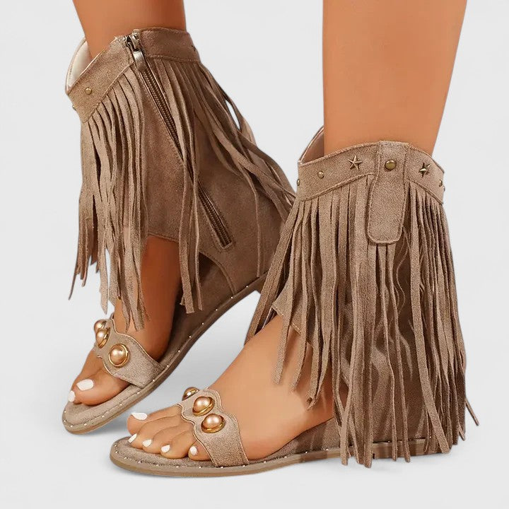 A woman wearing khaki fringe wedge sandals with a fringed ankle cuff and a back zip.
