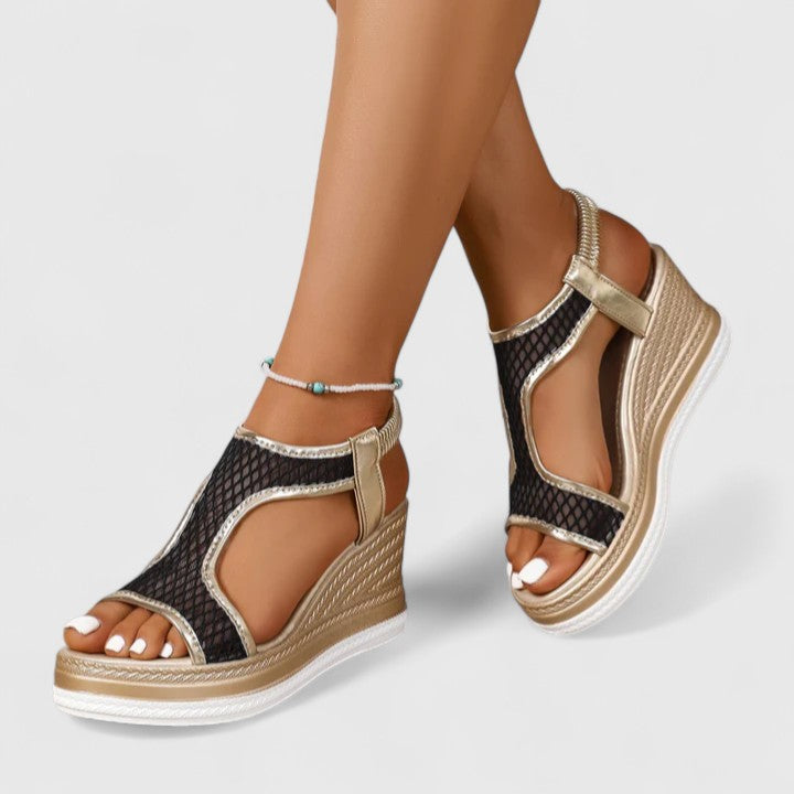 A woman wearing golden platform wedge sandals with a T-bar front, open-weave panels and a stretchy slingback.