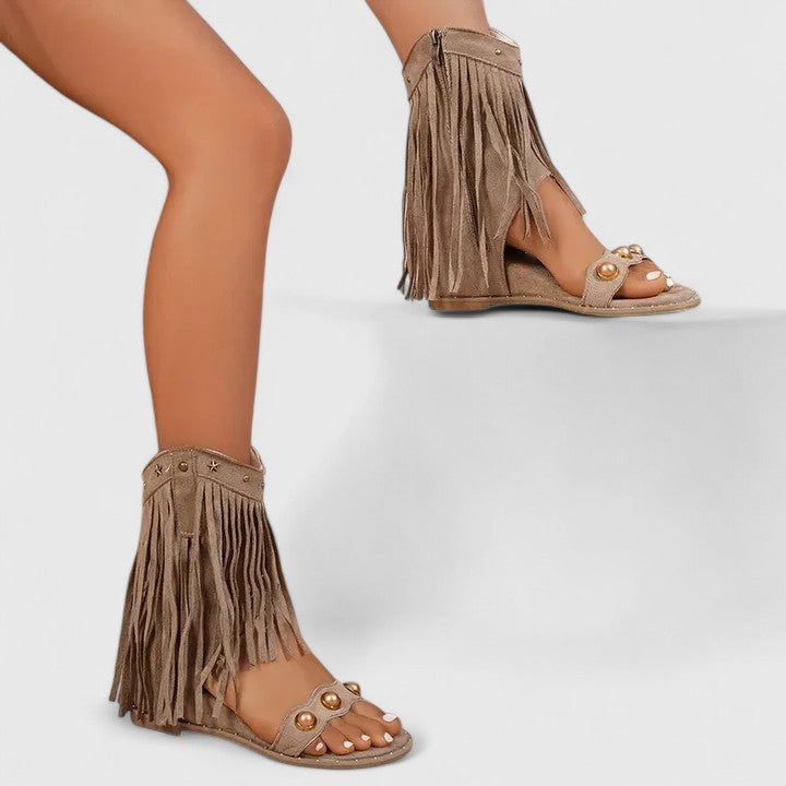 A woman wearing khaki fringe wedge sandals with a fringed ankle cuff and a back zip.