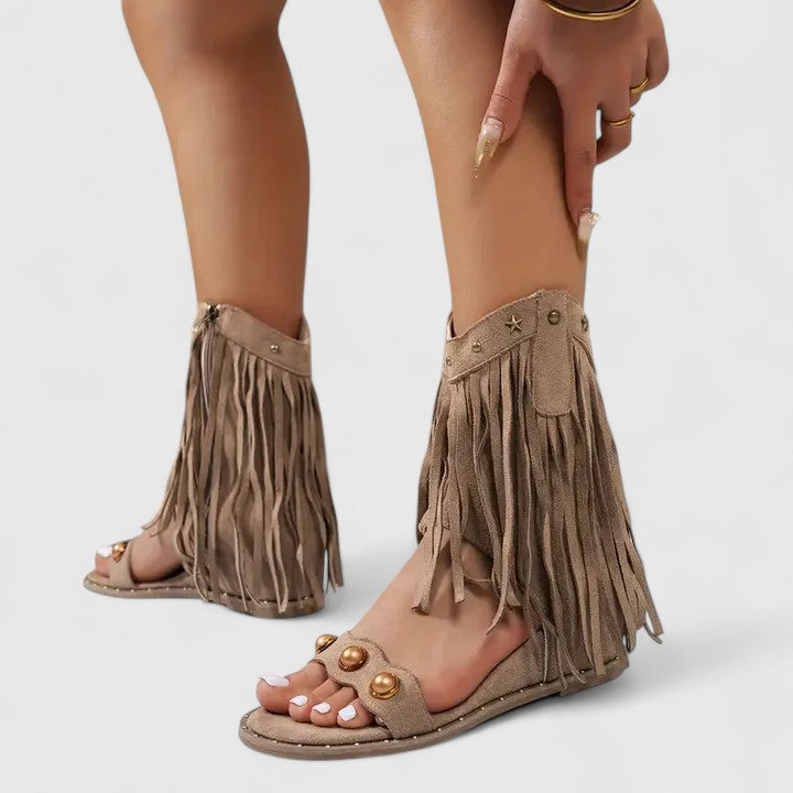 A woman wearing khaki fringe wedge sandals with a fringed ankle cuff and a back zip.