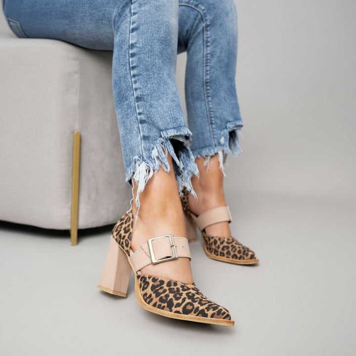 A woman wearing leopard print pointed-toe heels with a wide nude buckle strap and a neutral block heel.