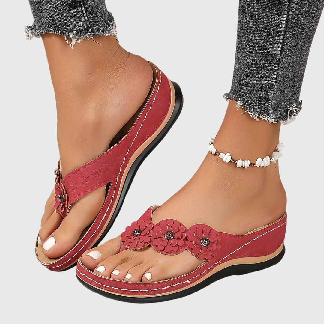 A woman wearing tan flower-trim flip-flops with a toe-post front and a low wedge sole.