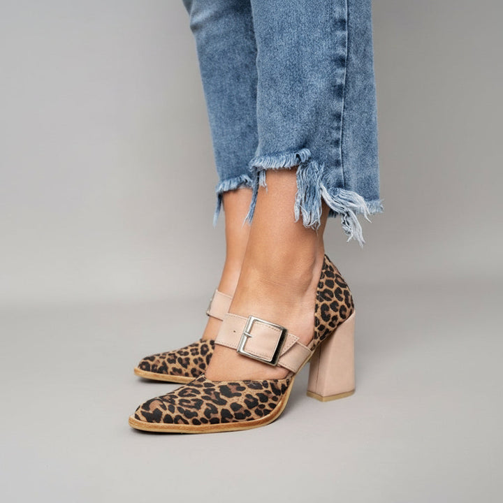 A woman wearing leopard print pointed-toe heels with a wide nude buckle strap and a neutral block heel.