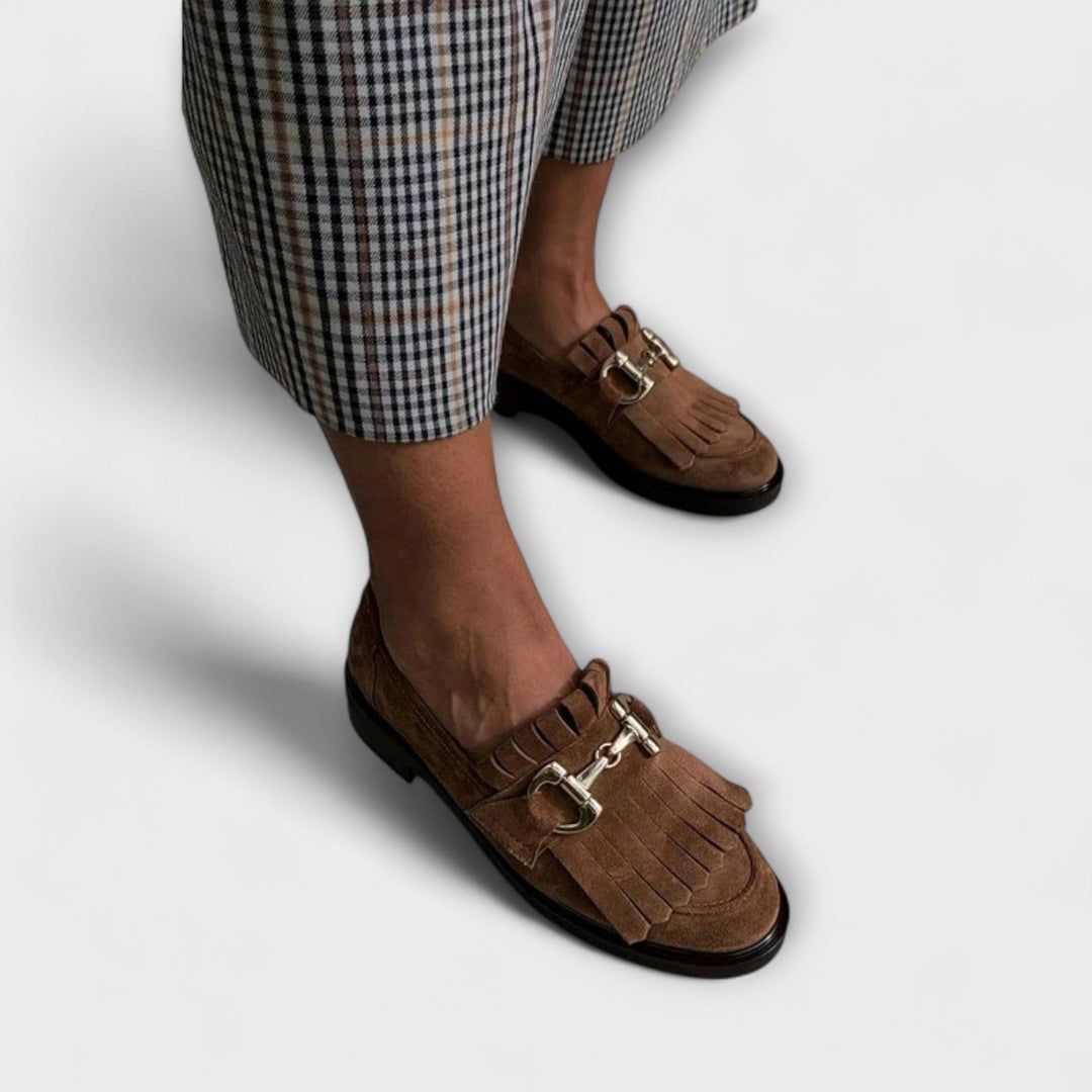 A woman wearing brown fringed loafers with a gold snaffle detail and checked pants, looking polished and at ease.