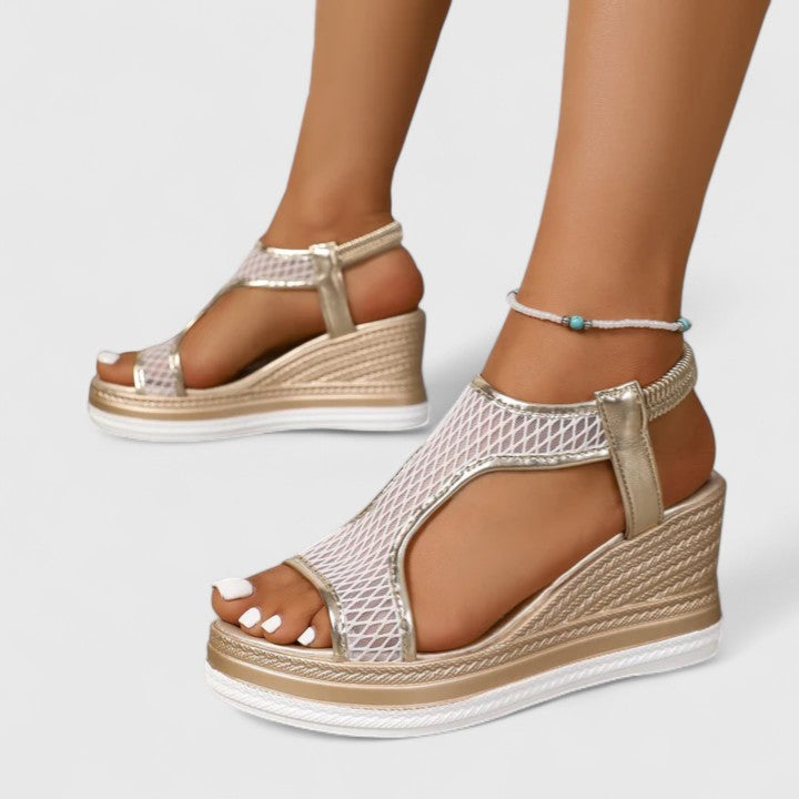 A woman wearing golden platform wedge sandals with a T-bar front, open-weave panels and a stretchy slingback.