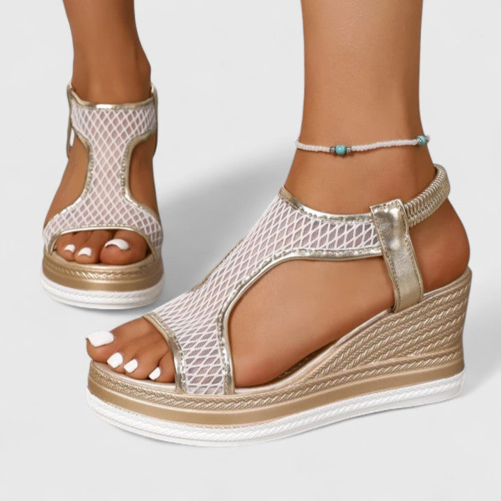 A woman wearing golden platform wedge sandals with a T-bar front, open-weave panels and a stretchy slingback.