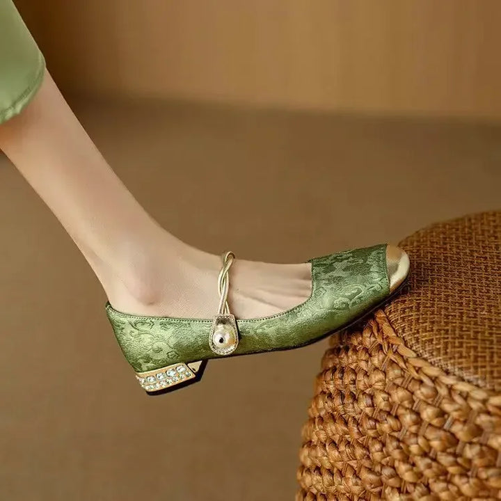 A woman wearing green low-heel Mary Janes with a twisted gold strap, gilded cap toe and a crystal-set heel