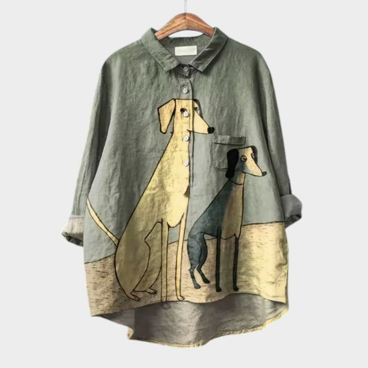 A relaxed blue button-up blouse printed with playful brown cartoon dachshunds and a small flower, shown on a hanger.