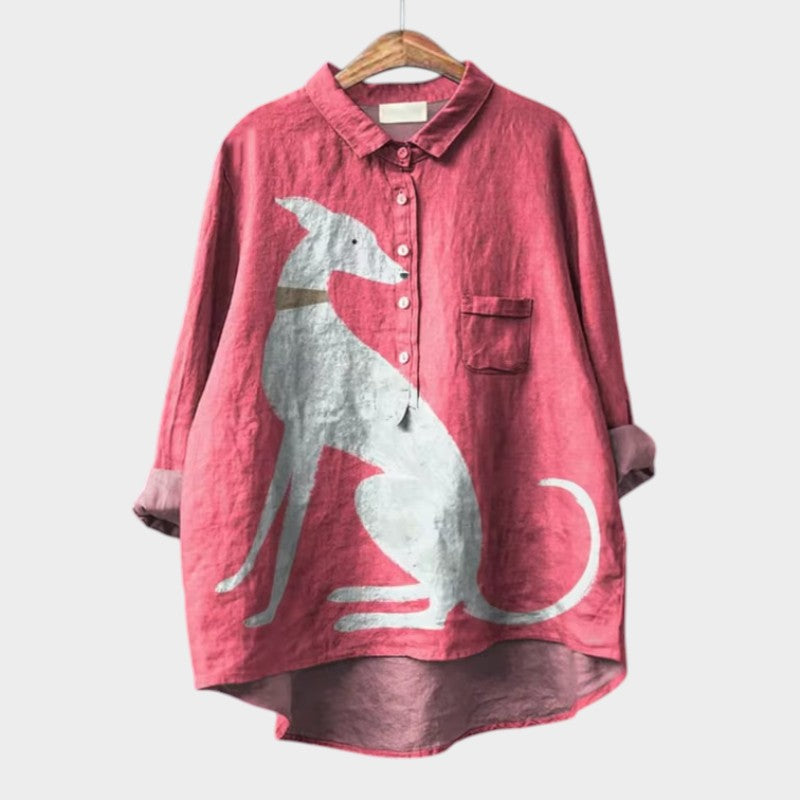 A relaxed blue button-up blouse printed with playful brown cartoon dachshunds and a small flower, shown on a hanger.