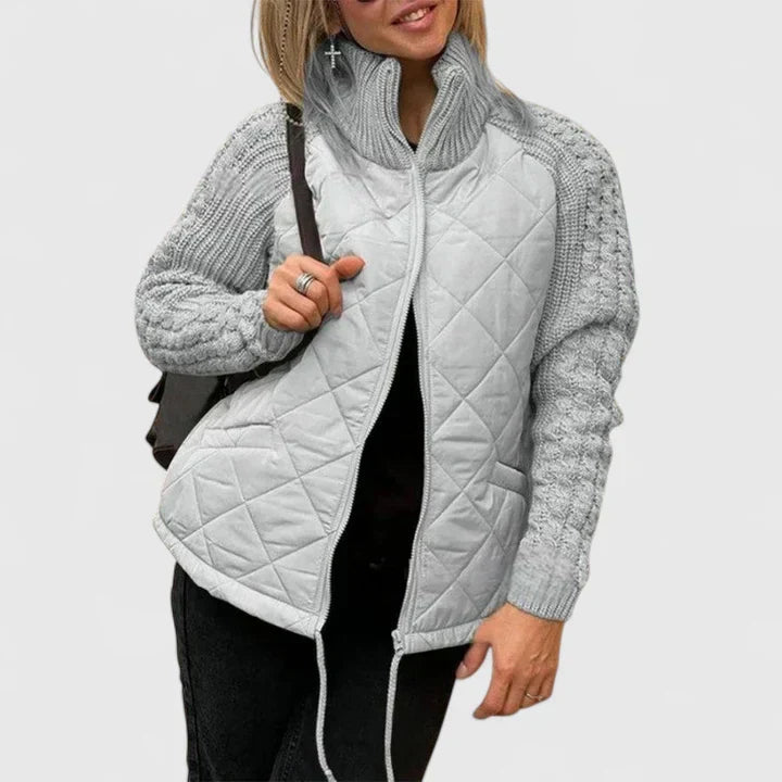 A woman wearing a light grey quilted jacket with chunky textured sleeves and a tall funnel collar.