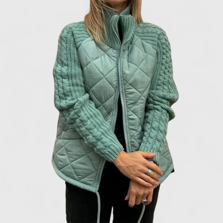 A woman wearing a light grey quilted jacket with chunky textured sleeves and a tall funnel collar.